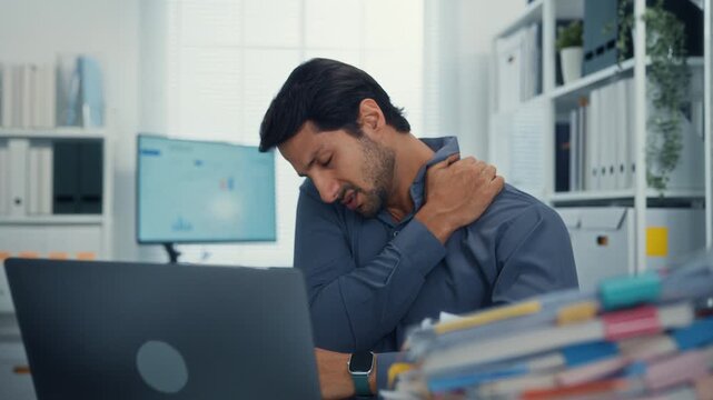 Young Latino businessman suffering shoulder pain and neck ache at office desk with computer. Office syndrome, muscle massage, overworked stress, bad posture, and health insurance injury concept.