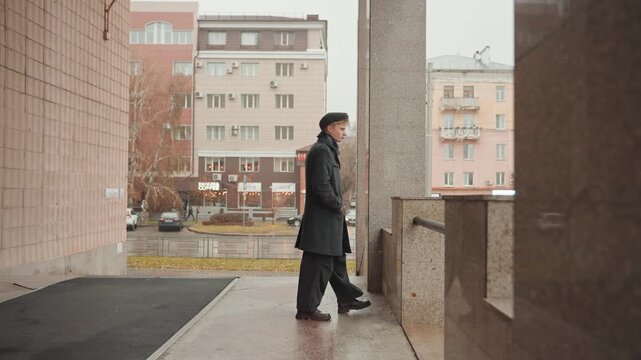 Fullbody white creator standing under archway, caucasian man in overcoat leaning on railing, urban entrance with granite columns, distant pastel buildings and rainy street, subtle motion as he waits,