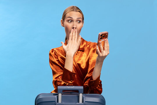 Surprised woman in orange blouse looks at smartphone with hand covering mouth. Blue background and suitcase in foreground. Concept of travel shock and unexpected news on mobile device.