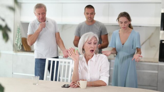 Woman sits at table in kitchen and pretends not to understand her family. High quality 4k footage