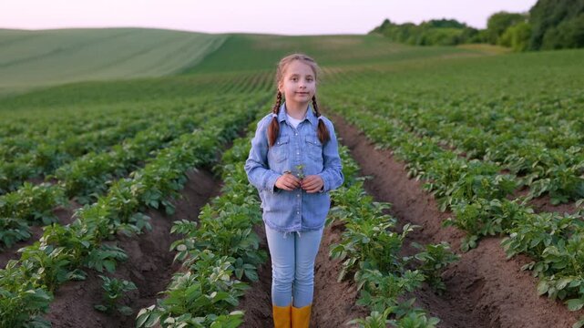 A smiling young girl with braided hair stands in a vast potato field wearing denim clothing