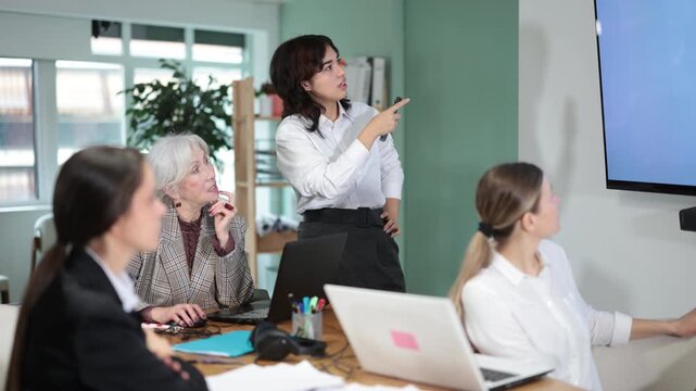 In study room, female business colleagues listens to lecture, look to presentation. Attendee remembers information during lecture, observes business coach. 