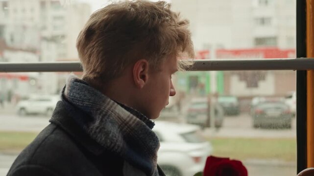Silent thinker looks through rainy window. Melancholy individual gazes beyond stormy city streets. Contemplative figure holds crimson flower while observing rainy urban environment quietly