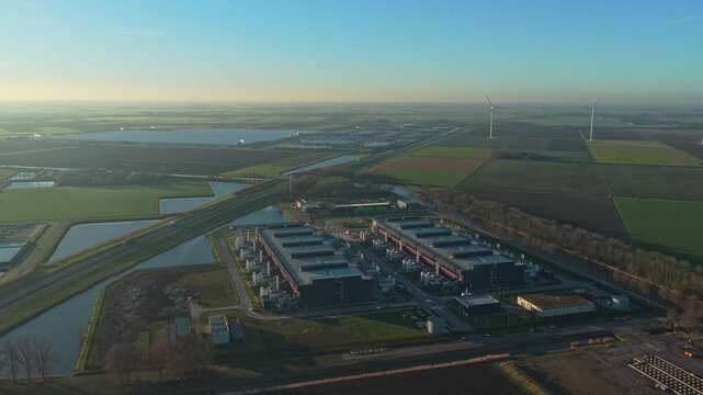High aerial perspective of a large data center complex surrounded by rural landscape. Modern infrastructure with wind turbines generating renewable energy