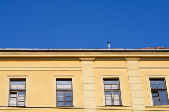 The yellow facade of a historic building features four rectangular wooden windows and decorative pilasters under a clear blue sky