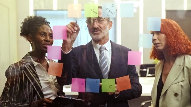 Diverse team collaborates at a glass board covered with colorful notes as a manager writes ideas during a strategy meeting in a modern office.