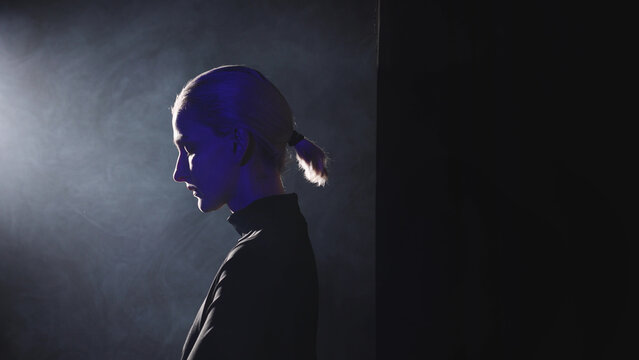 A young blonde woman with a short ponytail steps into a powerful side light. High-contrast video with her thoughtful profile illuminated, symbolizing the decision to stop being silent and embrace publ