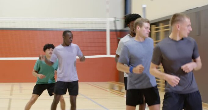 Diverse male teammates forming single-file jogging forward past volleyball net at gym for warmup