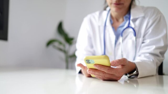 Female doctor with stethoscope using a smartphone for telehealth consultation, medical professional scrolling on a mobile app while sitting at a desk in her modern office