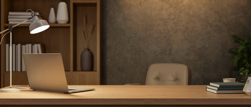 Laptop under lamp light on wooden counter table with beige leather chair and bookshelf in dim office