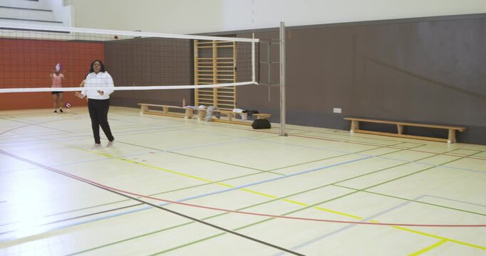 Diverse female volleyball team in school gym practicing drill, server tossing ball, coach directing