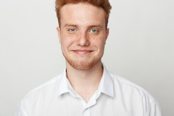 Obraz premium Smiling young man with red hair and beard wearing white shirt looks at camera on light background. Portrait of confident male with friendly expression and casual style.