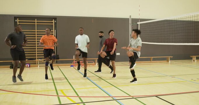 Male athletes warming up with high-knee hops in gym following grey-capped coach cues by net