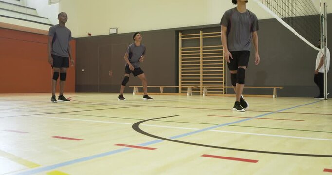 Male team in kneepads jumping reacting to incoming volleyball, passing at gym by net coach watching