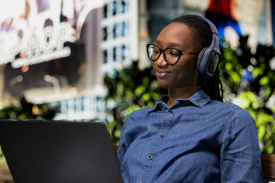 Relaxed person on park bench uses laptop to watch movies, smiling. Happy African american woman wearing headphones enjoying nice day outside, consuming entertainment media content on notebook