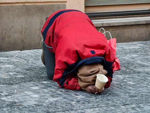 Person wearing red jacket kneels with head bowed on icy sideway in front of weathered wall