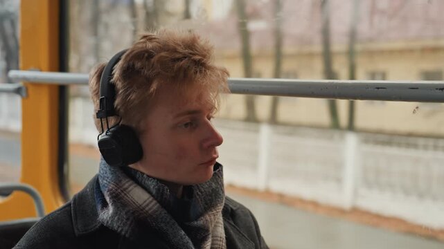 Young caucasian creator on tram listening to music through large black headphones, wrapped in scarf and coat, gazing out rainy window, pensive mood, aspiring composer sketching ideas on phone, soft
