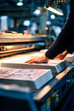 Workers use hands to operate an industrial paper machine in a printing house, producing printed materials. Posters are stacked on the table nearby