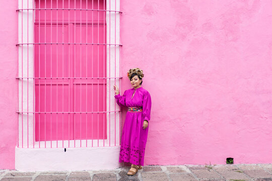 A woman in a bright pink traditional dress stands next to a pink building with a window with bars.
