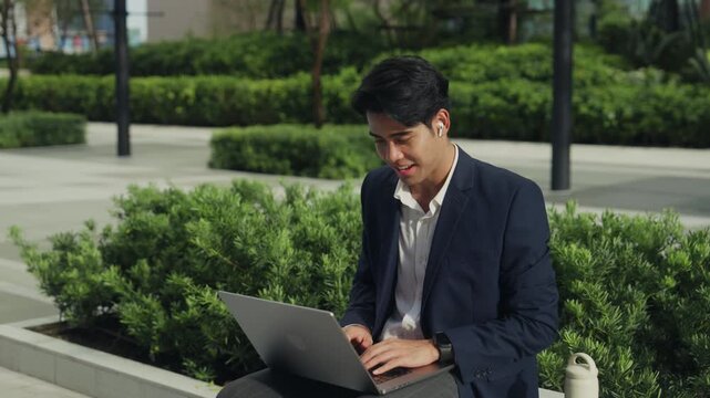 Young businessman sits in a city park, joining a virtual meeting on his laptop. Surrounded by nature and urban energy, he reflects hybrid work freedom, balancing flexibility and professionalism