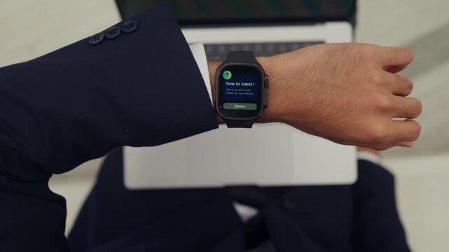 Young businessman closes his laptop after a smartwatch alert to stand and stretch, embracing wellness tech and mindful work habits, balancing productivity with health in a modern workday