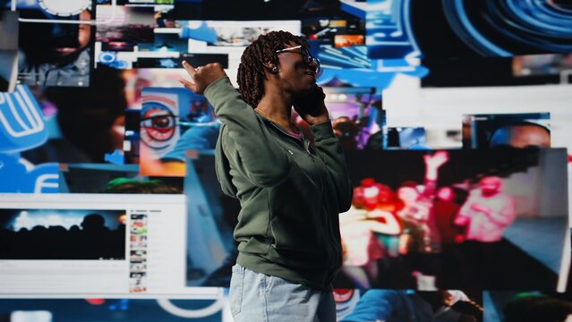 Smiling woman using smartphone to call friend, discussing movies, standing in front of vibrant video wall collage. Happy person excited to call mate on phone and chat about tv shows, camera A