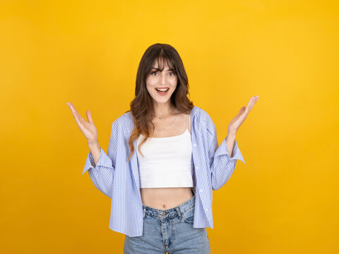 Excited caucasian woman raising hands with surprised expression, smiling widely while reacting, wearing striped shirt and denim, isolated on bright yellow studio background with copy space.