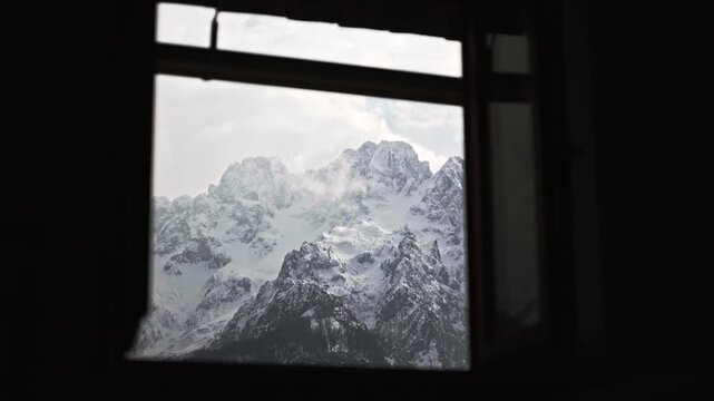 A beautiful scenic wide shot through the window of a cozy alpine cottage, looking out at the majestic, snow-capped peaks of the Julian Alps in Slovenia during winter.