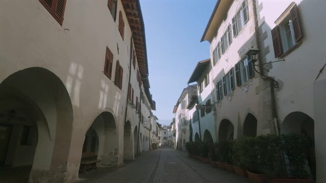A POV walking shot through the famous 14th-century stone arcades (Portici) of Egna, Italy. Captures the authentic medieval atmosphere and rustic merchant charm of one of Italy&rsquo;s mos