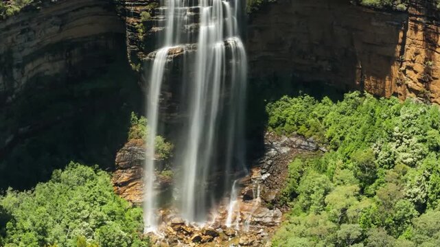 Time-lapse footage of a tall waterfall cascading down a steep cliff face into a rocky area surrounded by dense green vegetation. Multiple streams of water flow rapidly.