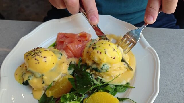 Close-up of hands cutting into eggs benedict dish with hollandaise sauce, smoked salmon, and fresh greens on a white plate in a bright dining setting