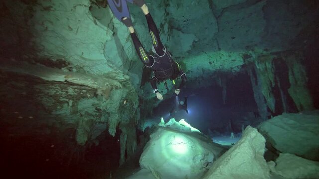 A scuba diver uses a handheld light to navigate a submerged cave chamber with rock formations and suspended particles in Cenote Dos Ojos, Mexico