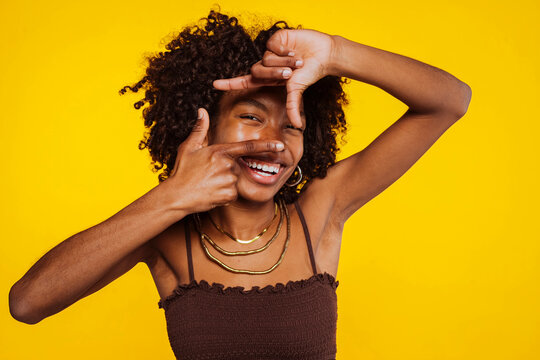 Joyful latin american black girl with afro hair smiling and making a finger frame at studio isolated over yellow background.