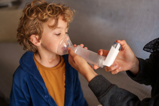 Child with curly hair receiving breathing treatment via an asthma inhaler and spacer, with an adult helping