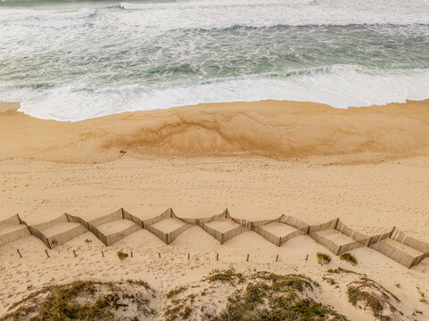 Aerial view of the beach with rhythmic wave patterns meeting the warm sand, and a geometric fence protecting the dunes, Quiaios Beach, Figueira da Foz, Portugal.