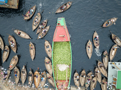 Aerial view of wooden boats clustered around a larger vessel filled with green produce, the water reflecting the sky above, Dhaka, Dhaka Division, Bangladesh.