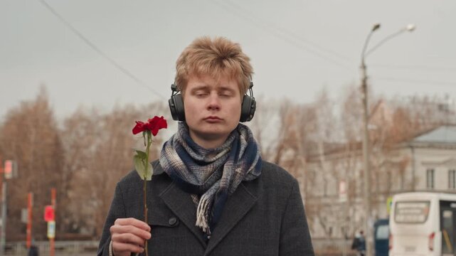 White man headphones holding red rose, quiet city square, overcast sky, wool scarf and coat neutral gaze shifts to ironic smirk and middle finger gesture, creator persona mixing romance and rebellion,