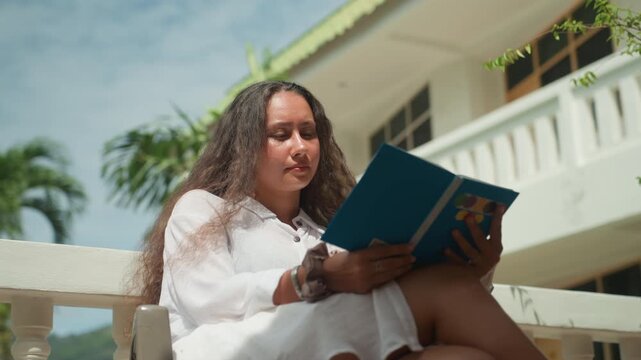 Woman reading outdoors. Woman in white enjoying sunny balcony read. Gentle breeze as woman studies travel guide outside. Peaceful vacation scene with woman reading guide beneath palm trees