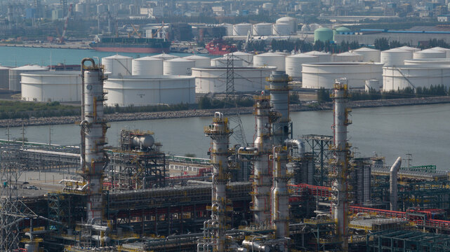 Aerial view of industrial complex with towering structures against the backdrop of white storage tanks and a distant cityscape, Tambon Map Ta Phut, Chang Wat Rayong, Thailand.
