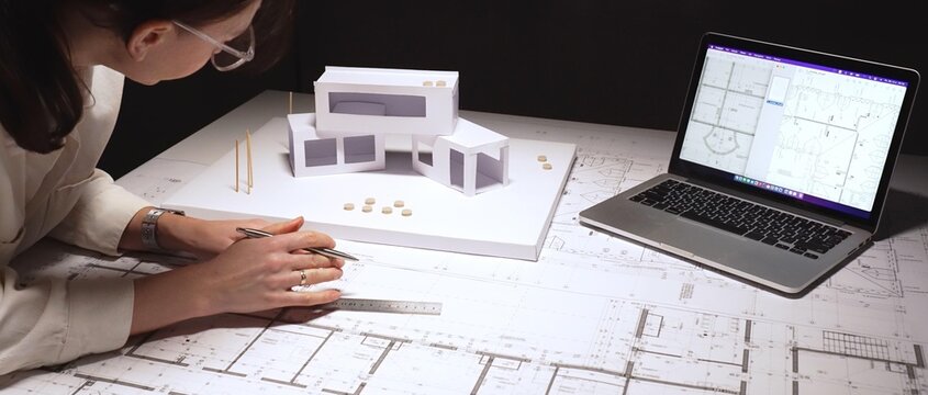 Architect carefully drafting detailed building blueprint plans, developing structural engineering designs with a laptop displaying cad software and a physical scale model on the desk