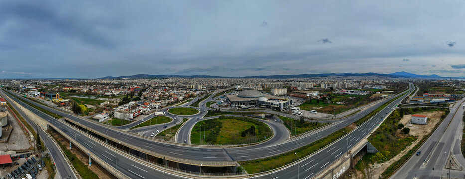 Aerial view of highways intersect near a modern building with a domed roof, cutting through the urban landscape under a cloudy sky, Thessaloniki, Thessaloniki, Greece.