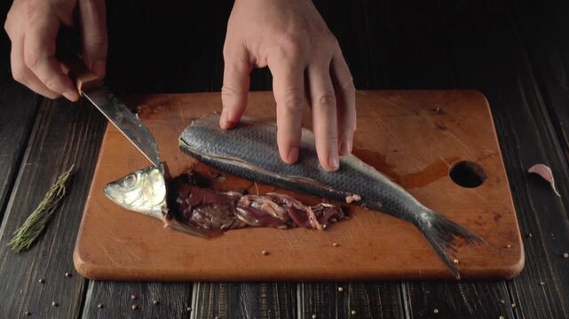 Hands preparing Atlantic herring fish on wooden cutting board, removing entrails and skin with knife, herbs and spices scattered around in a kitchen setting