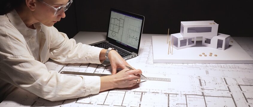 Adult female architect intensely working at a desk with blueprints, cad on laptop and a scale model, developing modern residential building plans and design solutions