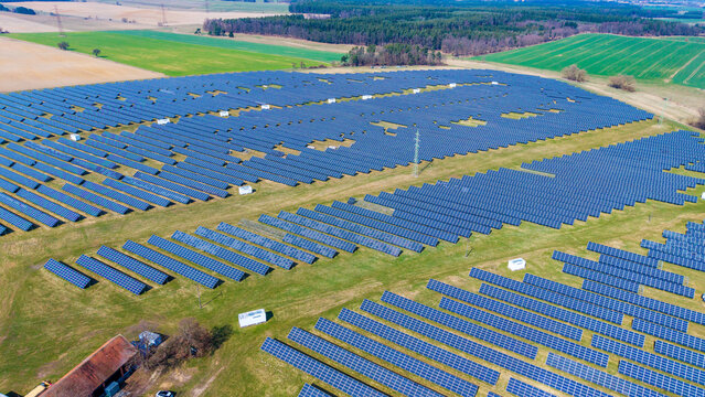 Aerial view of a vast solar array glinting under the sun, contrasting with the surrounding patchwork of green and brown fields, Sevetin, South Bohemian Region, Czechia.
