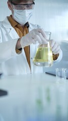 Fototapeta premium Male scientist wearing white protective gloves, mask and glasses is holding a yellow chemical solution inside an Erlenmeyer flask in a laboratory, vertical portrait. Science and medicine