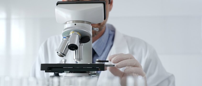 Scientific researcher wearing lab coat, glasses and white protective gloves carefully calibrating laboratory microscope, performing precision medical investigation. Medicine, healthcare and science