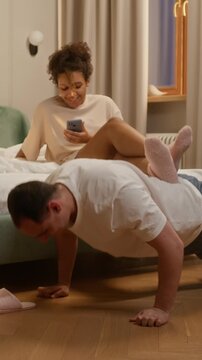 Vertical shot of African American woman sitting on bed, smiling and laughing while recording video of her husband doing push ups