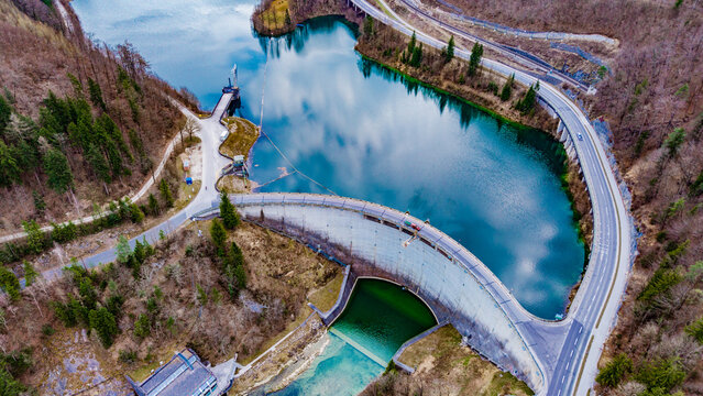 Aerial view of the Klaus dam holding back a serene lake reflecting the sky, embraced by roads and lush forests, Klaus an der Pyhrnbahn, Oberosterreich, Austria.