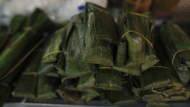 Stacked Banana Leaf Wrapped Rice Cakes On Metal Tray, Glossy Leaves Tied With String, Refrigerated Display Sheen, Readytoeat Snack, Bustling Evening Market Atmosphere, Savory Glutinous Texture, Seller