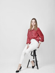 Fototapeta premium Junge Frau sitzt auf Hocker im Studio, elegante Pose mit roter Bluse und weißen Hosen, modernes Fashionporträt mit stilvollem Look und Copyspace